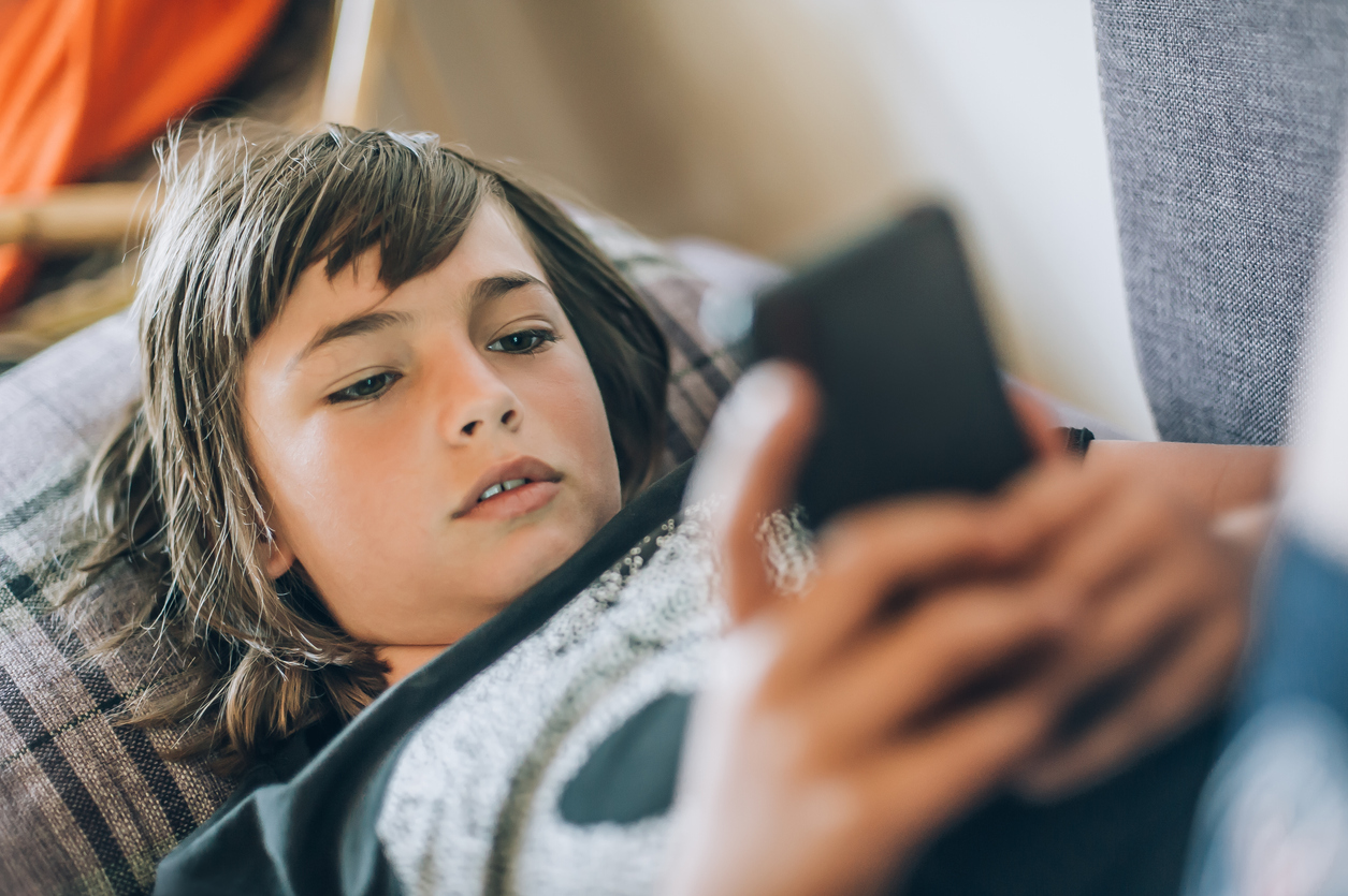 Teen boy checking social media holding smartphone, using mobile phone app playing game, relaxing on sofa at home. Emotional isolation and depression
